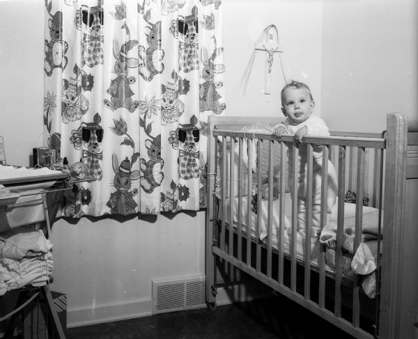8-month old Billy Gately of 4210 Nakoma Road standing in his crib in a nursery decorated with colorful stuffed animal print drapes.