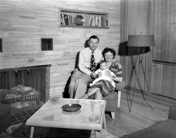Portrait of Ernst and Marie Friedlander with their baby, Helen, in the living room of their home that Ernst designed. The room includes a stone wall fireplace, built-in shelves, Mid-Century modern table and lamp, and large double-paned windows.