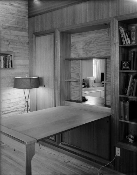 View of the dining room in the Ernst Friedlander house. The room features wood paneling, a dining table folded out of the wall, a view of the kitchen through the resulting wall opening, built-in bookshelves, and a Mid-Century modern lamp.