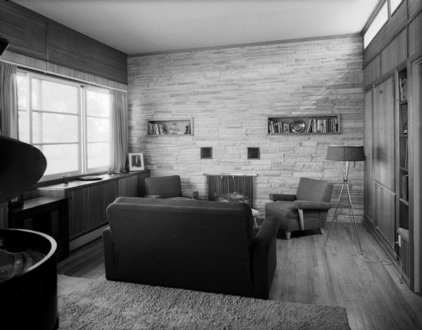 Interior view of the Mid-Century modern living room in the home of Ernst Friedlander. The room features a stone wall fireplace, built-in shelves, a fold-into-the-wall table, Mid-Century modern furniture and a double paned window.