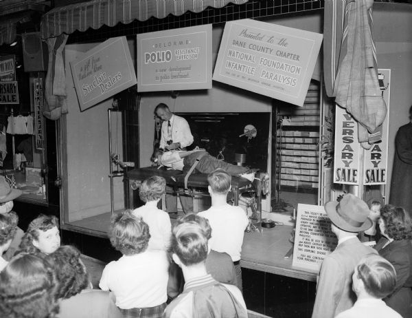 Crowds of shoppers watch the demonstration of a polio resistance exerciser held in a store display window. Dick Murphy, captain of the U.W. boxing team, is shown receiving "treatment" on the machine by Frank Lam of the Elgin Exerciser Appliance Company, makers of the machine. It will be used in polio treatment at Wisconsin General Hospital and was donated by Sinclair gasoline dealers to the Dane County Chapter of the National Foundation for Infantile Paralysis.