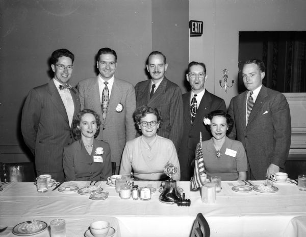 Group portrait of members of the Madison Exchange Club and their wives sitting and standing at the speakers table during their ladies' night banquet. Standing, left to right: James Oeland, program committee member; Harley Thronson, club president; Dr. Svend Riemer, the main speaker; Russ Gilbertson, committee member and Tom McGovern, program chairman. Seated in front, left to right: Betty Thronson, Mrs. Riemer, and Ruth McGovern.