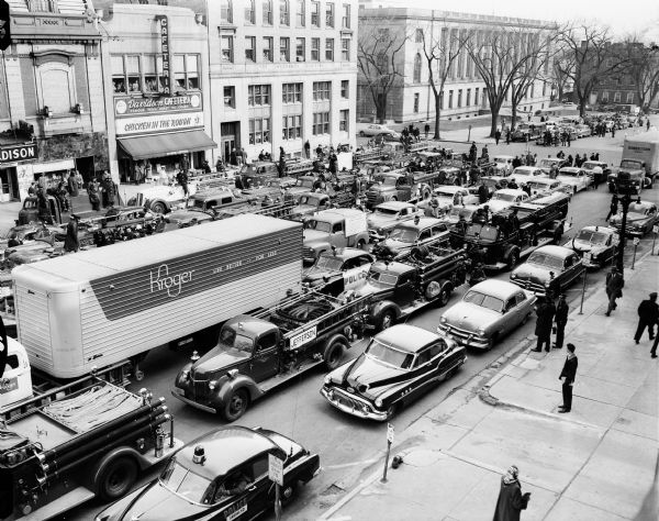 Elevated view of the 125 vehicles which were used in Wisconsin's first civil defense support battalion test in Madison. The vehicles were parked on Monona Avenue in front of the State Capitol. Businesses on Monona Avenue are Madison Theatre (113), The Perfume Shop (115), Davidson's Cafeteria (117), Beaver Insurance Building (119), and the Federal Building/Post Office (215).