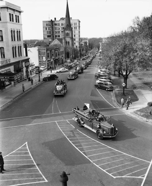Elevated view of a parade of vehicles for Wisconsin's first civil defense support battalion test in Madison approaching along South Carroll Street and turning onto West Main Street at the Capitol Square. The marquee of the Park Hotel is in the foreground and Grace Episcopal Church is in the background.