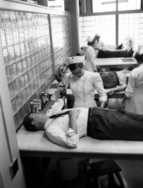 Alderman Lyle R. Andrews, 7th ward, donates blood while lying on a gurney as Red Cross staff nurse Marge Schmitz looks on. Two nurses and other donors can be seen in the background.