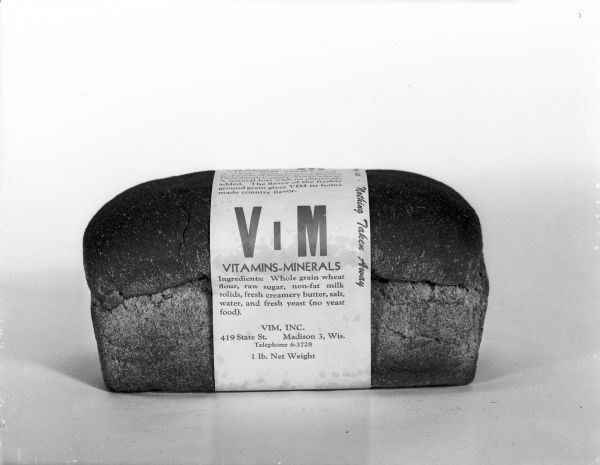 Advertising studio photograph of a one pound loaf of whole wheat bread wrapped in a paper label.  The label reads, Vim bread vitamins and minerals made with whole grain wheat. The bread was baked by Vim, Inc. at 419 State Street in Madison, Wisconsin.