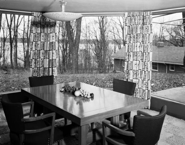 Interior view of dining room in Stewart and Jean Douglas home at 811 Farwell Drive. The view includes Mid Century table, chairs and draperies, and two floor-to-ceiling windows joined together in a seamed invisible corner. Beyond the windows are a descending bluff and lake, and a building on the right.