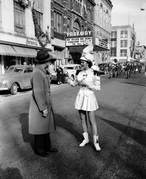 Governor Walter Kohler and Sonja Iverson, Cambridge High School drum majorette, standing on West Mifflin Street during the Cambridge scrap metal parade around the Capitol Square. Parkway Theater is in the background. Proceeds from sale of metal will go towards a movie projector for Cambridge High School.