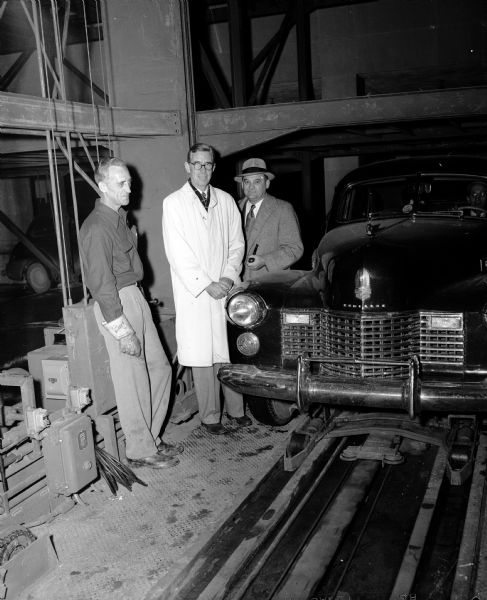 The first commercial pigeon hole parking unit in the world was built for the Harry S. Manchester department store, behind Manchesters at the corner of Wisconsin Avenue and East Dayton Street. Shown, left to right, are: inventor Leo Sanders of Spokane, Washington; Morgan Manchester, president of the store, and Mayor George Forster. Also shows a car with its wheels raised and its front axle resting on a tracked moving mechanism. The Manchester pigeonhole parking unit was six stories high and it held 169 cars. "The parking is all done mechanically, without a hand touching the auto after the driver steps out. The hydraulically-operated lift moves sideways and vertically and can park 168 cars at an average time of 35 seconds per car when operating at peak efficiency."