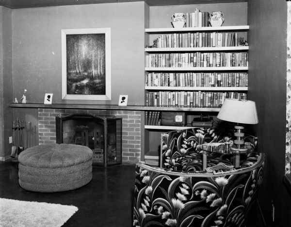 Living room in the David & Florence Bade residence at 5701 Winnequah Road in Frost Woods, Monona. A large ottoman stands in front of the brick fireplace. Two pieces of the three-piece sofa are arranged around a round end table. The wall next to the fireplace is made up of built-in bookshelves.