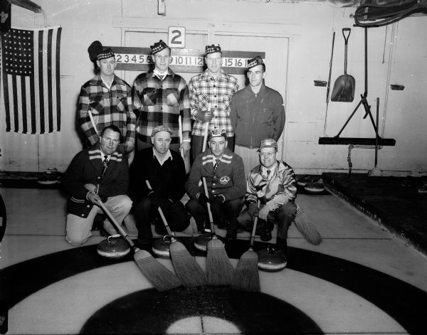 Group portrait of men at the Reynolds rink of Lodi, taken at the annual Midwest Curling Association bonspiel held in Madison. Pictured are, left to right in front, Hugh Reynolds, Bob Bilke, Hub Noltmeyer, and Jim Schmitt. In the back, left to right, are Russ Nugent, John Woody, Deane Woody, and LaVerne Harmon.