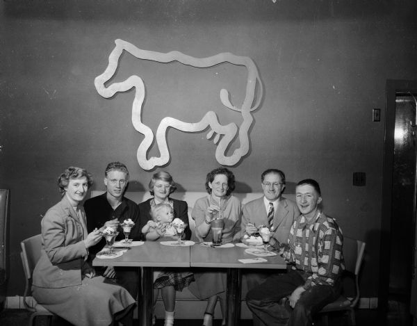 Gorup portrait of the Russell E. Frost family seated in a ice cream booth, eating a variety of frozen confections. Left to right: Sue Frost; Jack Frost; Betty Bancroft Frost, holding infant daughter, Lynn Marie; Mrs. and Rusell Frost; and Billy Frost. Photograph was used for the family Christmas greeting card.