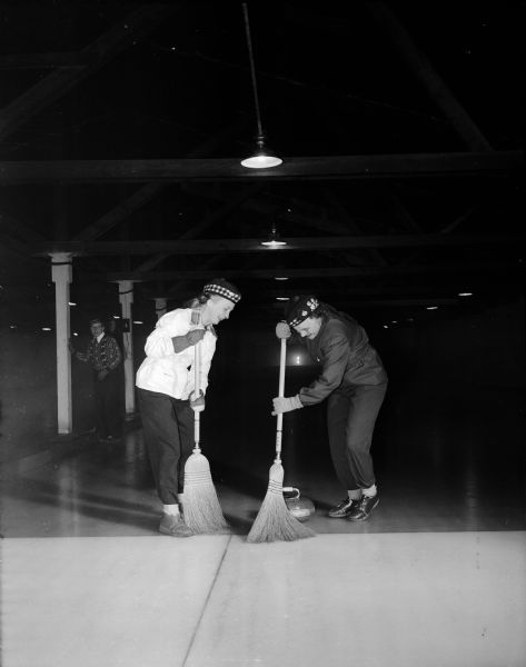 Mrs. Stephen Resan of Route 1 (left) and Agnes Gurney "sweep a stone" on a curling rink. The women are members of the Madison Toories, a women's curling group.