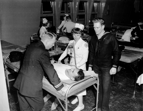 Navy Radioman Burton Fischer is shown on a cot as he donates blood during a blood drive for naval reservists from the naval reserve training center. At left is Lieut. James McIntosh, a physician. The Red Cross staff nurse at center is Erika Brechler, and on the right is Hospitalman William Gilardi.
