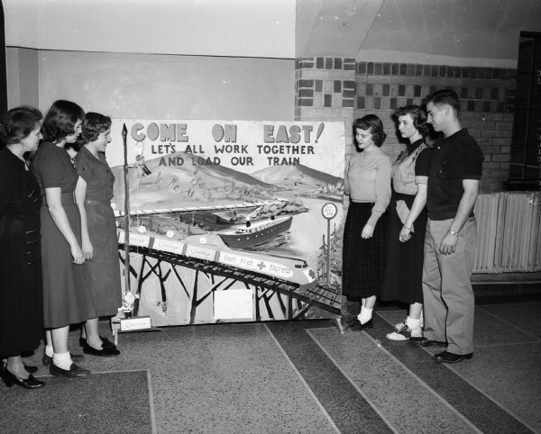 Members of the East High School Junior Red Cross Gift Box Drive committee gather around one of the posters that urged student participation. From left are: Ethel Davey, the faculty advisor, and students Donna Hubanks, Myrna Ballinger, Joan Bradley, Florencia Roh, and Charles Deadman. The boxes collected contained useful items to be distributed to sick and needy persons.