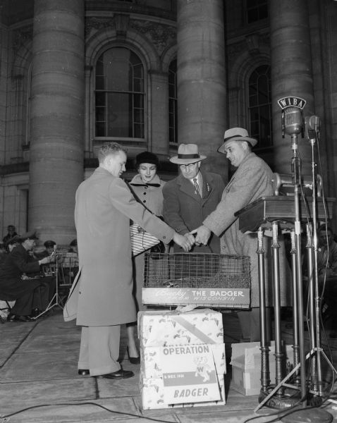 Pvt. Gregory Becker, Beaver Dam (left), leader of the forty-seven "Operation Badger" Air Force recruits, is presented with numerous gifts to present (including a badger in a cage) to San Antonio and Texas officials when the group arrives at the Air Force base there. Presenting three gifts are Miss Geri Robinson, Miss Recruiter for Wisconsin; A.E. Wegner, representative of Governor Kohler; and Mayor George Forster.