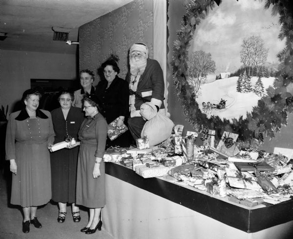 Members of the Business and Professional Women's Club stand beside a display of the many gifts to be donated to various organizations providing care for children. Standing at left in the foregound: Mrs. Virginia Sullivan, assistant youth counselor at the Wisconsin School for Girls in Oregon, one of three organizations to receive gifts; Mrs. Hilda Engroff, and Mrs. Sadie Herr, members of the club's social committee. Standing in the rear are Mrs. Charles E. (Alice) Hemingway, club president; Miss Blance Hartman, member of the social committee; and Miss Louis Morrisey (Santa Claus).