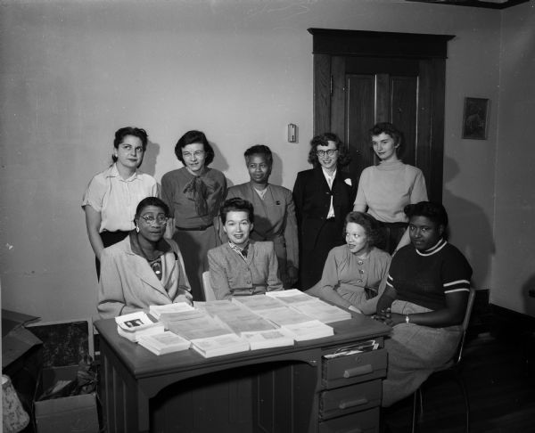 Portrait of nine members of the Friendship Club, meeting to work on a cookbook. The Friendship Club is an interracial women's club, formed to develop mutual understanding among themselves to answer the problem of segregation-ignorance. Sitting, left to right: Ola Jordan, Evelyn Kolman, Gladys Tomczyk. and Mrs. Jerome Parks. Standing, left to right: Mrs. Malcom Gordon, Mrs. William Cook, Gertrude Taliaferro, Alicia Ashman and Judith Gutman. 
