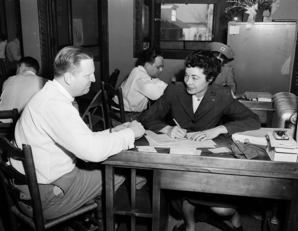 District Attorney Richard W. Bardwell, seated at left, is shown registering with Mrs. Walter J. (Charlotte) Kohler, the governor's wife and a Red Cross worker, during the annual Red Cross blood bank drive. In the background is Assistant District Attorney Roland R. Day registering with another Red Cross worker.