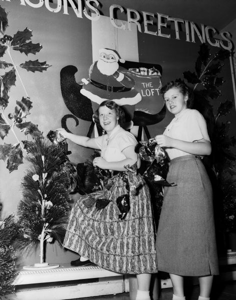 Two teenage girls are placing a garland on an artificial Christmas tree in front of a graphic depicting Santa in a sleigh holding a bag of presents. They are Carolyn Ames and Sharon Thompson and they are decorating at the Loft, a teen center.