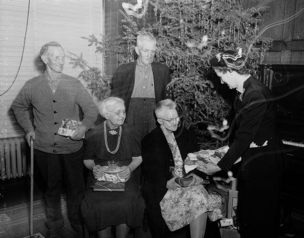 Residents of the Dane County Home for the Aged in Verona are pictured while receiving gifts and lunch at the Madison Association of Underwriters' annual Christmas party. Left to right: Martin Eassel, Julia Simonson, Cleo Wallace, Grace Sommers, and standing at far right serving cookies and coffee, Emily Widen, association member.