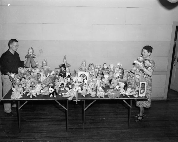 Individuals standing beside a table laden with a collection of one hundred sixty dolls and stuffed animals collected at a square dance for the <i>Wisconsin State Journal's</i> Empty Stocking Club. The dance was sponsored by the Nichols and Allis square dance groups. In the photograph are Lloyd Heim and Mrs. F.R. (Alice) Shores who helped set up the "store."