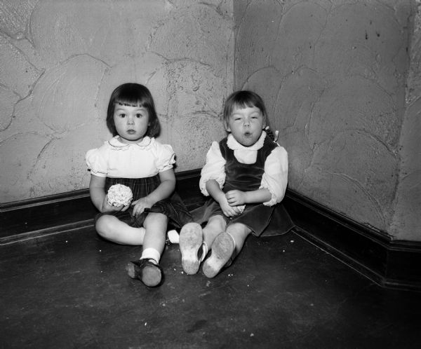 Two children sitting on the floor in a corner are posing for a portrait at the annual Christmas party for children and grandchildren of University Club members. At left is Anne Harris, holding a popcorn ball.
