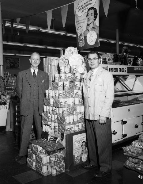 Two unidentified men stand next to a display announcing a .01 cent sale of Old Dutch cleanser in a Lancaster grocery store. A banner annoucing the product for sale hangs above them.