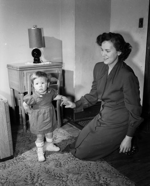 20-month-old Beverly Smith standing while wearing braces on her lower legs and holding the finger of her mother, Mrs. Eugene Smith.