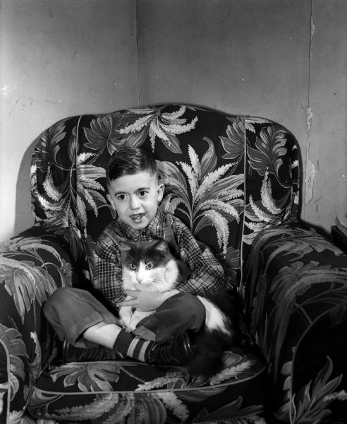 Warren Anderson, age 7, former polio sufferer, is shown sitting in an upholstered chair with his cat, Sam. Warren was saved by his 14-year-old sister, Carol, who noticed the boys neck was stiff. Within hours he was being treated for polio in a Madison hospital where it was also discovered he had spinal meningitis germs.