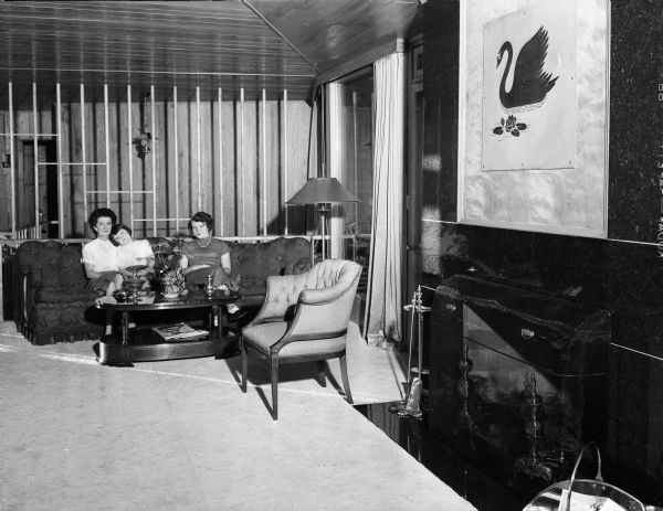 Mrs. John Booher and her two daughters sitting on a sofa in the first floor living room of their home overlooking the Baraboo river near Reedsburg. The poles behind them hold up the hanging staircase that leads to the ground floor.