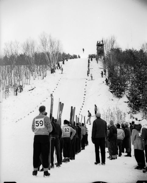 Bob Leeman of Minneapolis heads off the ski slide at Tomahawk Ridge near Middleton into the "wild blue yonder" as skiers and fans watch below. No. 59 is identified as Gilbert Ross of the Blackhawk Ski Club, sponsors of the annual meet. Ahead of him among the spectators is another skier, No. 82, Phil Brockington of Beloit.