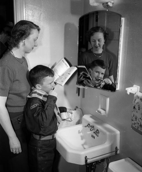 Cub Scout Marshall R. Hanks, Jr. brushes his teeth at a bathroom sink, with mother standing by checking directions for tooth brushing in a Cub Scout book.