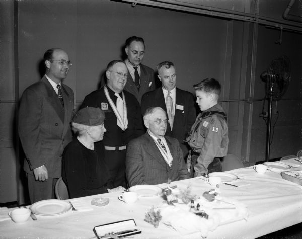 One of the newest members of the Four Lakes Boy Scout Drum and Bugle Corps, John Wilbur, chats with Marvin B. Rosenberry, former chief justice of the State Supreme Court. Also pictured, standing in the rear from left to right, are: Ralph D.L. Price, president of the Corps' parent organization; C.H. Beebe, executive director of the Corps; Pete H. Norg, Four Lakes council executive; and Norman Critser, president of the Madison Optimist Club, sponsor of the Corps. Mrs. Rosenberry is shown seated to the left of her husband.
