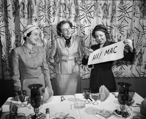 For the first time in the history of the Madison Advertising Club, the women members are planning the dinner and program to be held at the Hoffman House. Hats made from materials used in their various types of advertising will be worn by the female members attending the dinner and program. Shown making arrangements for the dinner are, left to right: Merle O'Rourke, treasurer (the only position in the organization open to women), Badger Lane; Peg Thom, 24 Sherman Terrace, and Charlotte Peterson, 109 W. Doty Street, general chairman of the program. Peterson's poster displays the official slogan of the club, "Hi ! MAC." Other women assisting in the plans for the dinner are Frieda Brockhausen, Ferne Noreen, and Mrs. Norman Gill.