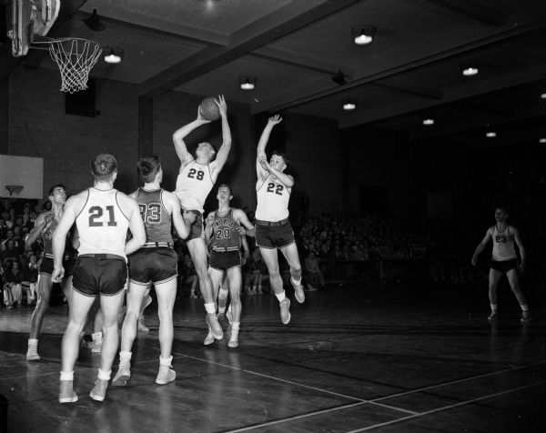Charlie Brendler (white #28), Madison East center, and leading scorer in the Big Eight Conference is shown airborne, with the ball about to sink a rebound shot. Other East players in white are Jim Kurth (21) and Ted Blackney (22). Central players are Norbert Schachte (33), Loren Tipler (20), and Gib Pond under the basket.