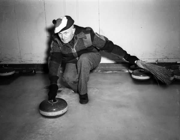 H.T. Ferguson, veteran Madison curler, squatting with his hand on the curling stone handle, about to propel it. He is wearing a tam on his head. The club bought 66 new "stones" for almost $2,000. The granite to make the "stones" is fetched up from the bottom of the ocean off Scotland. The sea salt action makes the rock especially tough.