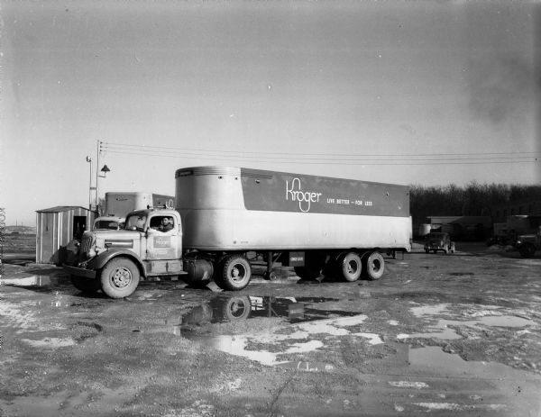 A Kroger grocery store chain semi-trailer truck parked at 2701 Packers Avenue with a man in the driver's seat. The sign on the side of the truck reads: "Kroger Live Better -- For Less". Trucks and automobiles are parked near a building with loading docks in the background on the right.