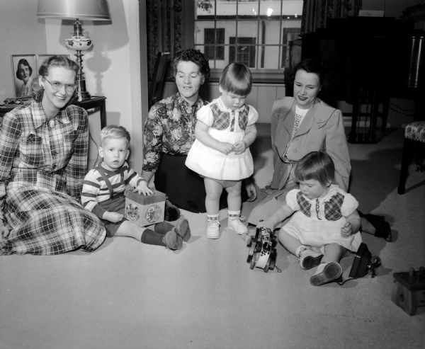 The junior division of the University of Wisconsin Service League for faculty wives has a number of interest groups including that of "Understanding Children," taught by Mrs. Leslie A. (Dora) Osborn, center, 4217 Wanetah Trail. Mrs. Osborn is pictured conducting a session of the relative merits of various types of toys. Her "guinea pigs" are Eric Anderson, 2, at left with his mother, Mrs. Laurens Anderson, 13-B University housing (whose husband is an assistant professor at the university), and Kathy and Karen Schmitiz, fifteen-month-old twins of Mr. and Mrs. Norbert (Ruth) Schmitz, 126 Bellevue Court. Mrs. Schmitz is pictured at right.