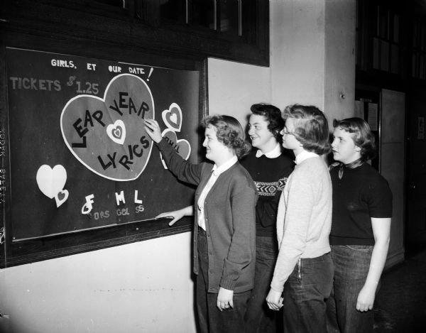 Four girls admire a poster advertising the Wisconsin High School "Lovers Leap" Dance.  From left to right are: Carol Jacobson, Peggy Neesvig, Gloria Jacobson, and Nancy Gunderson. Also shows the interior doors and windows in a hallway at Wisconsin High School.