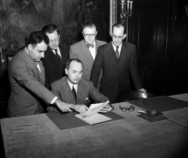 Governor Walter Kohler, Jr. seated at a desk in the governor's office with four men standing by. The photograph was taken during a Great Lakes-St. Lawrence Association meeting. Left to right: N.R. Danielian, Washington D. C., executive vice-president of the association; Robert W. Hansen, Milwakee, past national president of the Eagles; Frank H. Ranney, chairman of the Milwaukee Harbor Commission; and Curtis Hatch, Madison, president of the Farm Bureau Federation.