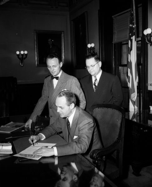 Two men watch Governor Walter J. Kohler sign a proclamation in his office, possibly for Heart Week.