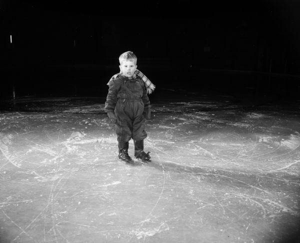 Johnny Beck, age 2, stands firmly and solemnly on his tiny double-runner skates as he watches the older Madison Skating Club skaters at Truax skating rink.
