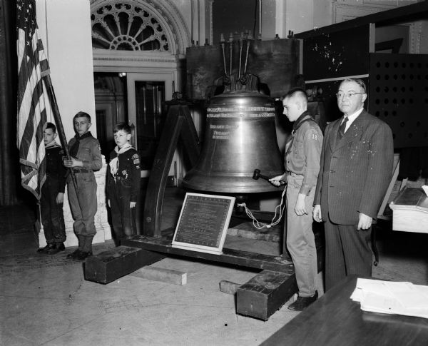 Boy Scouts ring a replica of the Liberty Bell to launch the three year progam of Wisconsin Boy Scouts. The bell stands in the Wisconsin State Historical Society Museum. Each of about 50 scout units from five Madison area counties endorsed the program. Scout David Lewis of Madison's Troop 18 tolled the bell. Members of the color guard shown at left of the bell are: Robert Mikulak of Cub Pack 302, Bobby Bulloch of Scout Troop 19, and Niels Nielsoen of Cub Pack 302. Scout David Lewis is pictured second from the right, and at the right is master of ceremonies, Justice Grover Broadfoot of the State Supreme Court.