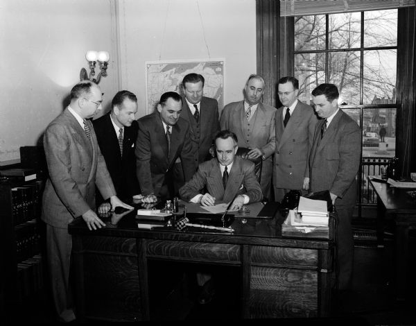 Wisconsin Attorney General Vernon W. Thompson sitting at his desk while signing an endorsement of the Madison observance of Crime Prevention Week. The national observance of the week is sponsored by the Exchange Club, and the Madison Chapter is in charge of plans for the area. Witnessing the signing, left to right, are: Delmar Schreier, chairman of the Crime Prevention Week committee; Fred Ferguson, club president; Madison mayor George Forster; District Attorney Richard W. Bardwell; Philip Oakey, Madison police inspector; United States attorney Thomas E. Fairchild; and John Brickhouse, committee member.
