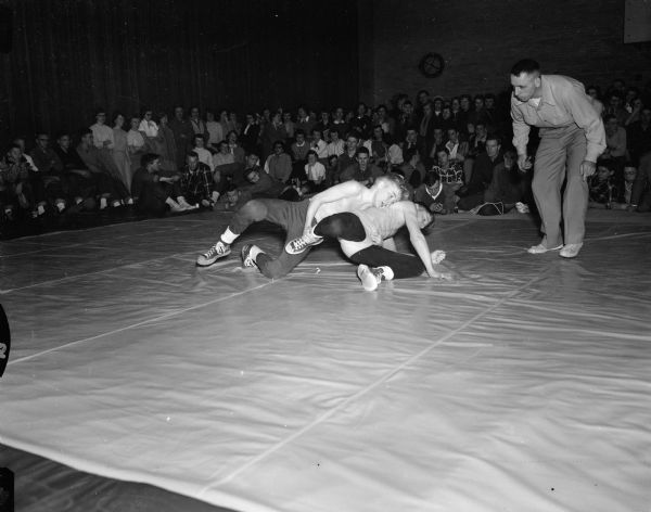 Two wrestlers grapple down on the mat as a referee looks on. An audience sits on the floor or stands in the background.