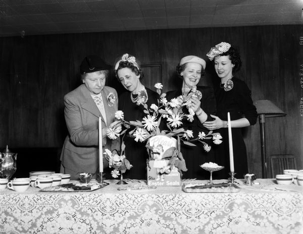 Four ladies are admiring the centerpiece at the tea held at the Memorial Union in honor of the members of the University League who volunteered to address envelopes for the Easter Seal Campaign. From left are Mrs. Edwin B. (Rosa) Fred, wife of the University of Wisconsin president; Mrs. Herman H. (Lorraine) Brockhaus, general chairman of the Easter Seal sale; Mrs. L.H. (Mildred) Adolfson, University League senior division; and Mrs. Warren C. Young, University League junior division.