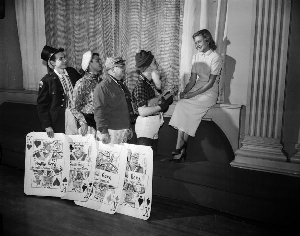 Seated at right is Phyllis Berg, a finalist in the University of Wisconsin Prom Queen Contest.  As part of her campaign theme, "The King and I," four of her costumed YMCA supporters hold large king playing cards. The supporters are, left to right, Sandy Smehoff, Al Menza, Jerry Marr, and Milton Erdman.