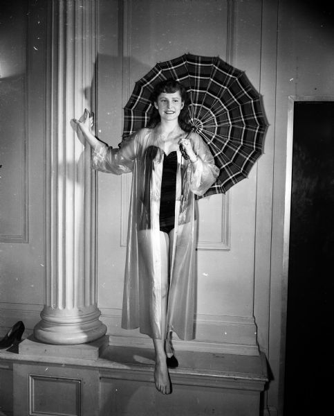 University of Wisconsin Prom Queen contest finalist Nancy Smith is shown wearing a bathing suit, and transparent rain coat, and holding an umbrella to portray her campaign motto: "If She Reigns, She'll Shine."