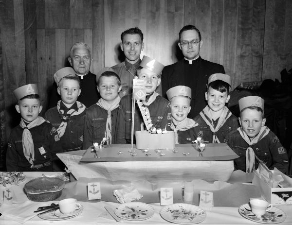 Members of Cub Scout Pack 315, Den 4, at Our Lady Queen of Peace Catholic Church are shown at their annual Blue and Gold Banquet. They are standing behind the model paper cardboard aircraft carrier they built to commemorate the 42nd anniversary of scouting. The Cub Scouts, first row, left to right, are: Steve Banaszak, Michael Morgan, Jim Shipe, Tom Edgerton, Michael Kessenich, and Jim Edgerton. In the back row, left to right, are: the Reverend F. L. McDonnell, pastor of the church; Arthur Kessenich, cubmaster, and the Reverend William DeBock, assistant pastor. They are all in uniforms, and the boys are also wearing paper sailor hats.